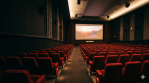 Interior of a cinema theatre with rows of red seats facing a lit screen showing a film